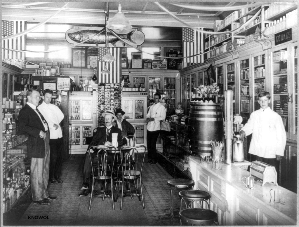Photograph shows the interior of a soda fountain/drugstore with customers and employees in Islip, NY in 1915.