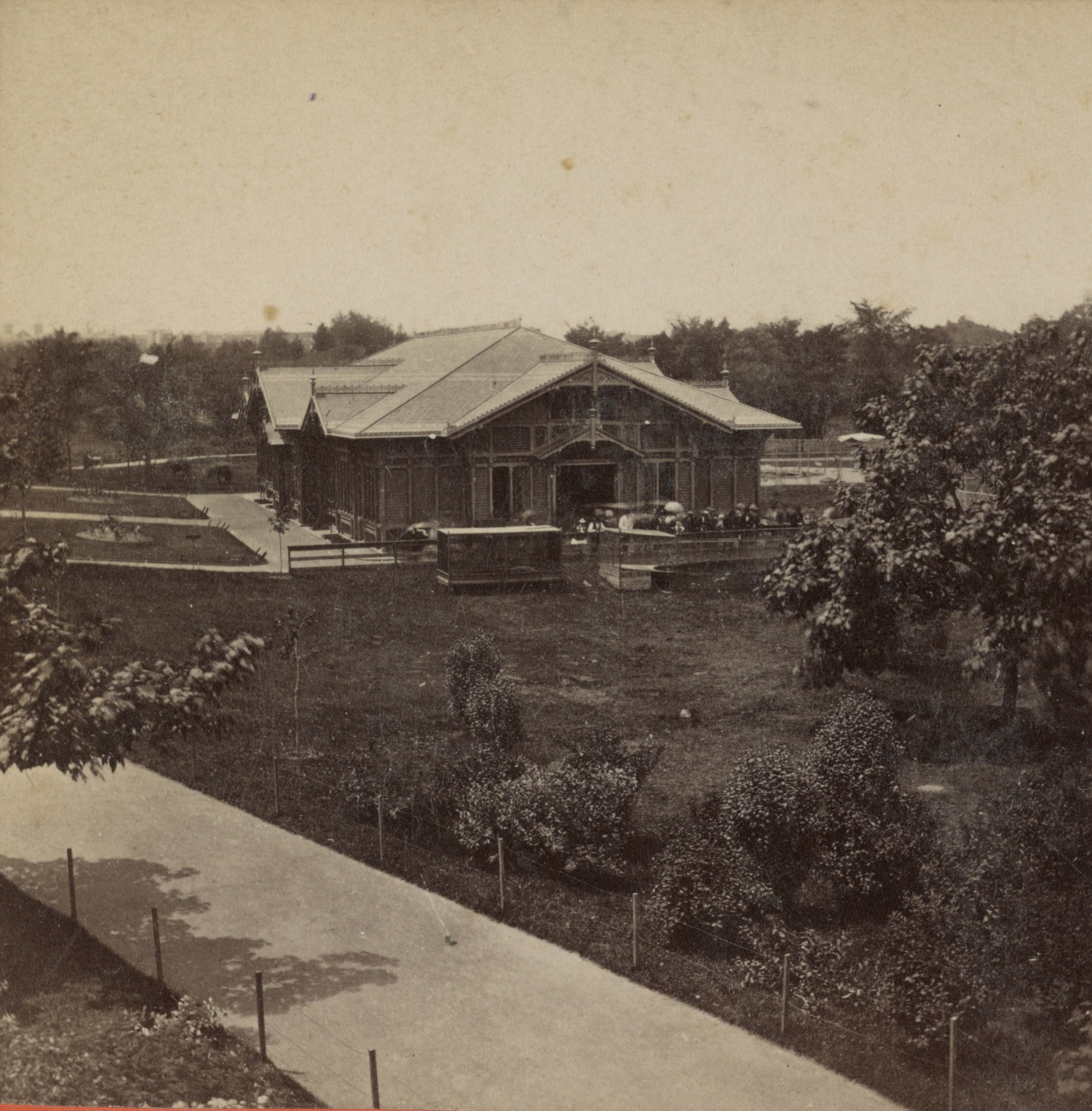 1870s photograph of the Menagerie building in Central Park, a one-story wooden hall with a peaked roof, lawns and animal cages in front