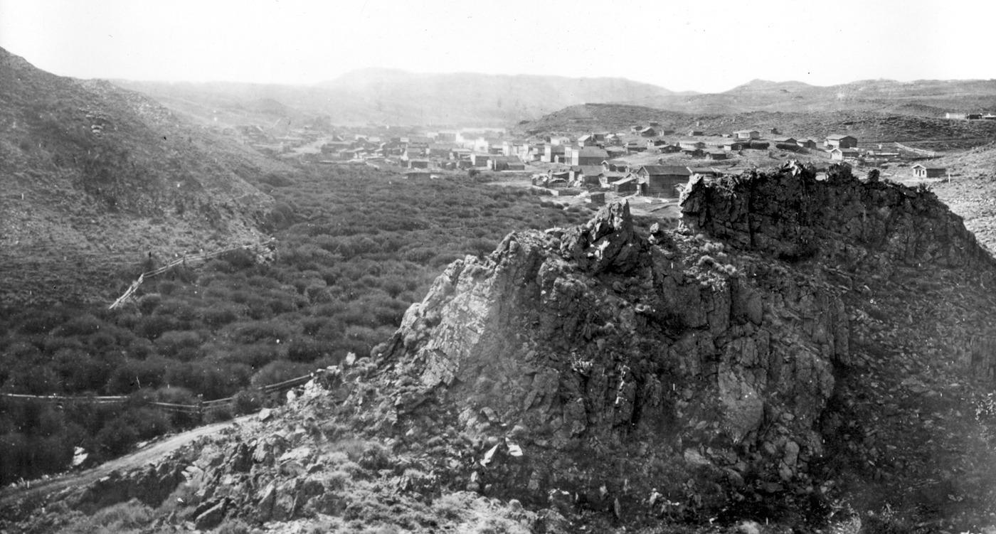 1870 photo of South Pass City, Wyoming, a mining camp of frame buildings in a valley of sagebrush hills, seen past a rocky outcrop