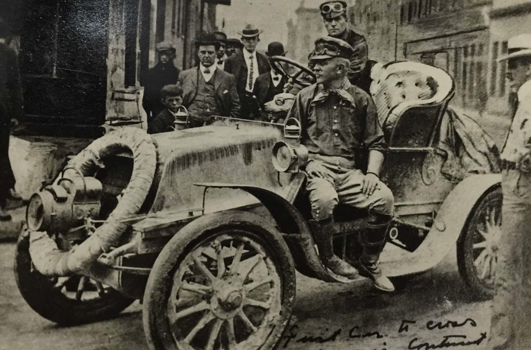 1903 photograph of a mud-covered open Winton automobile on a town street: a man in a cap and riding boots sits sideways on the car, a second man in goggles stands behind him, Bud the bulldog in goggles looks out by the steering wheel, and men in suits and hats watch from the sidewalk