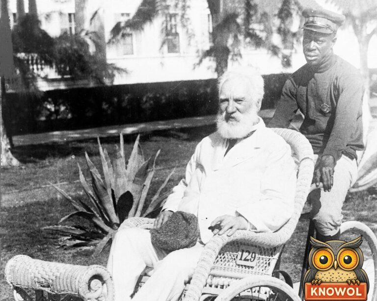 Alexander Graham Bell, white-bearded and dressed in white, sits in a wicker wheeled chair with a uniformed attendant on the bicycle seat behind him