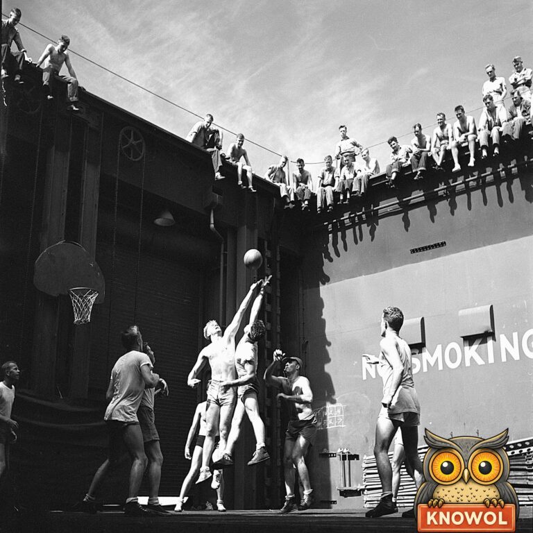 Navy officers in shorts play basketball in the well of an aircraft carrier's elevator, two men leaping for the ball while sailors watch from the deck edge above