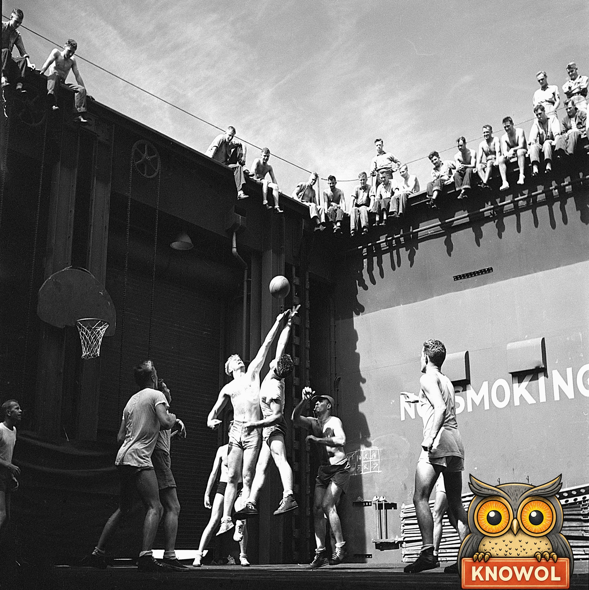 Navy officers in shorts play basketball in the well of an aircraft carrier's elevator, two men leaping for the ball while sailors watch from the deck edge above