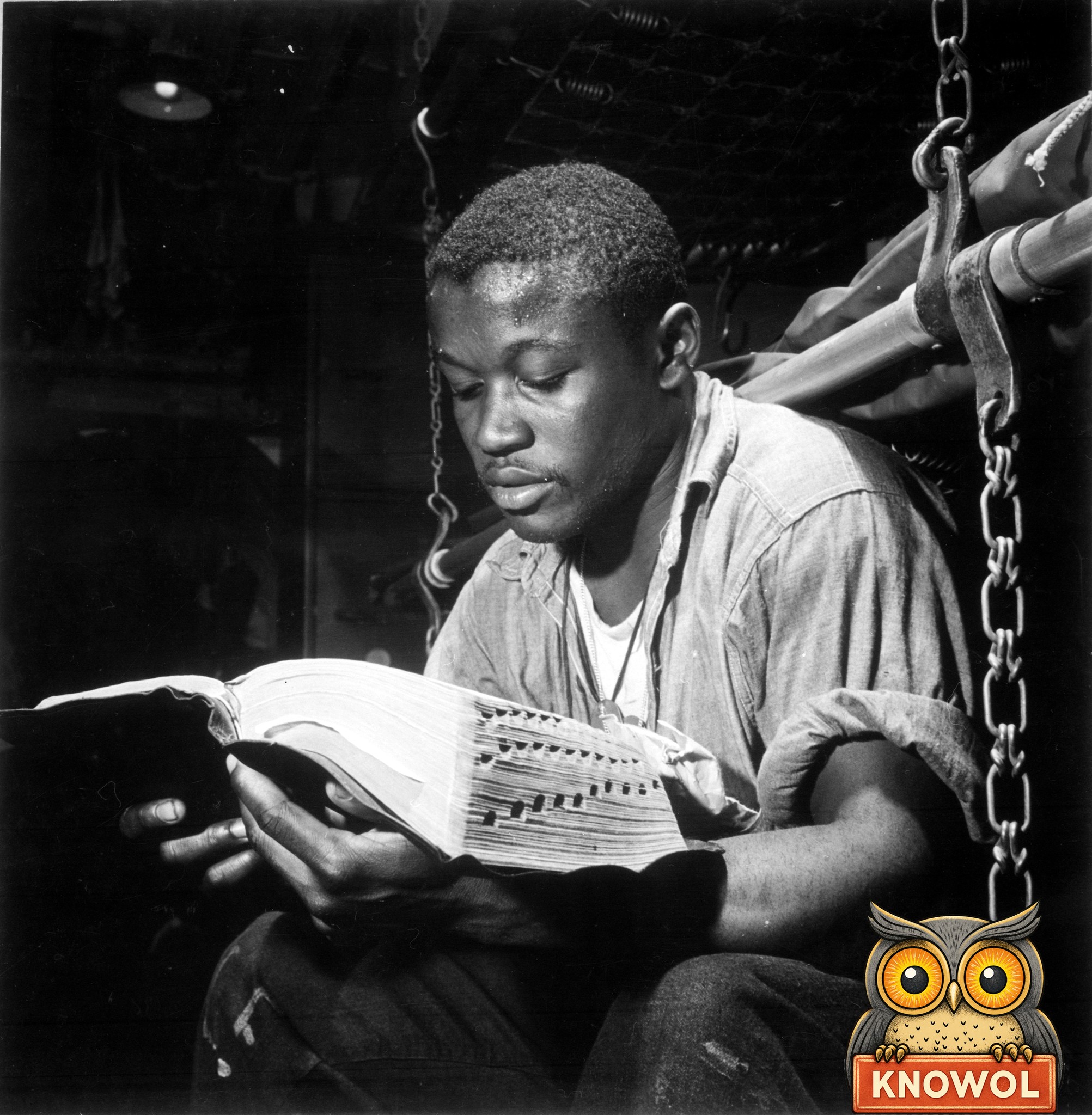 A young Black Navy sailor in a work shirt sits between chain-hung bunks below decks, reading a worn Bible by lamplight