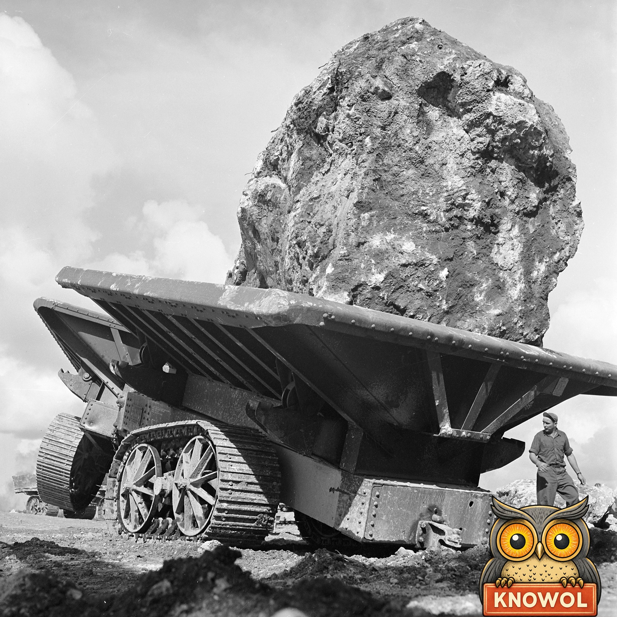 A huge rough boulder rides on a steel crawler wagon under a cloudy sky, with a Seabee in a cap standing beside the wagon's tracks