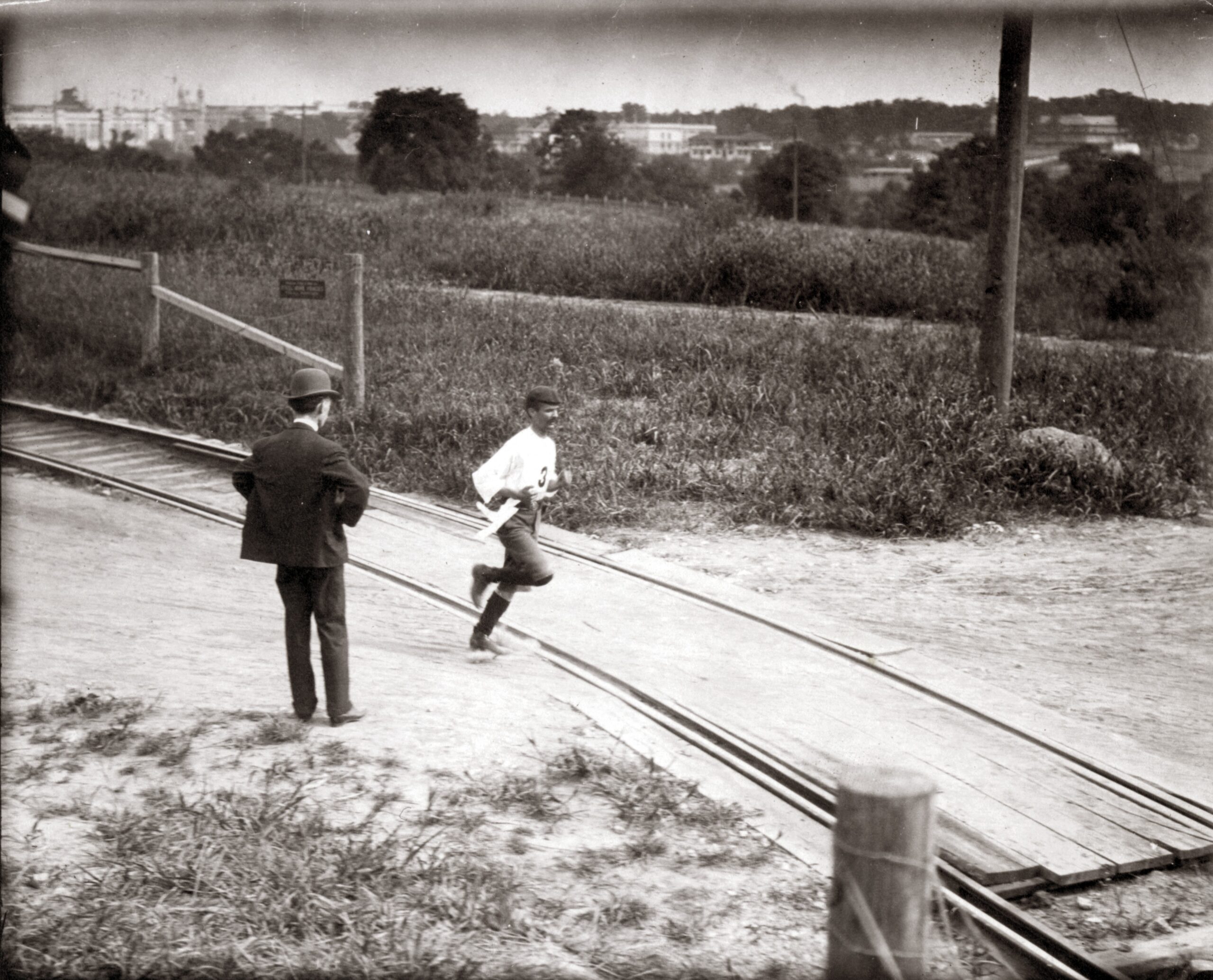 1904 photograph of Cuban runner Felix Carvajal in a white shirt, cap and cut-off trousers running across railroad tracks past a man in a bowler hat