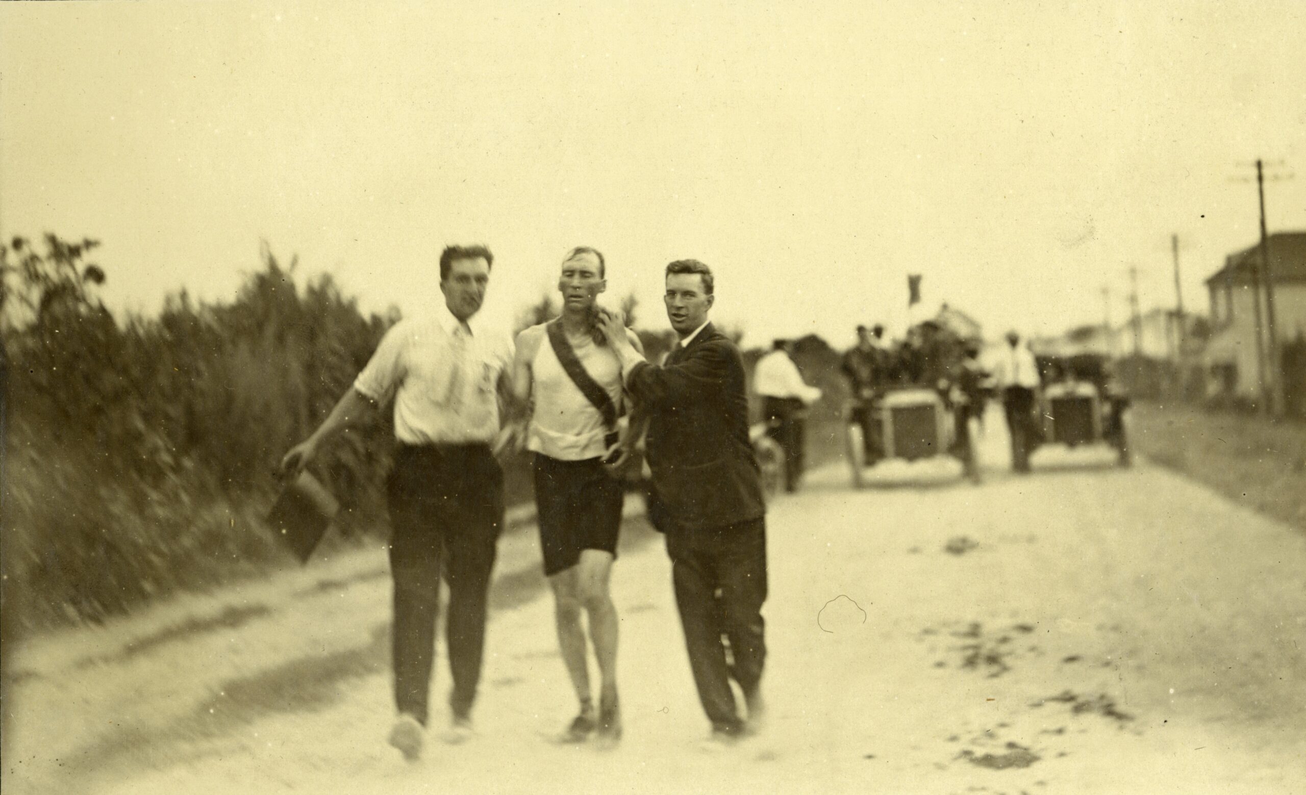 1904 photograph of marathon winner Thomas Hicks, in a white running shirt and dark shorts, held up by two men in street clothes on a dusty road, with early automobiles following behind