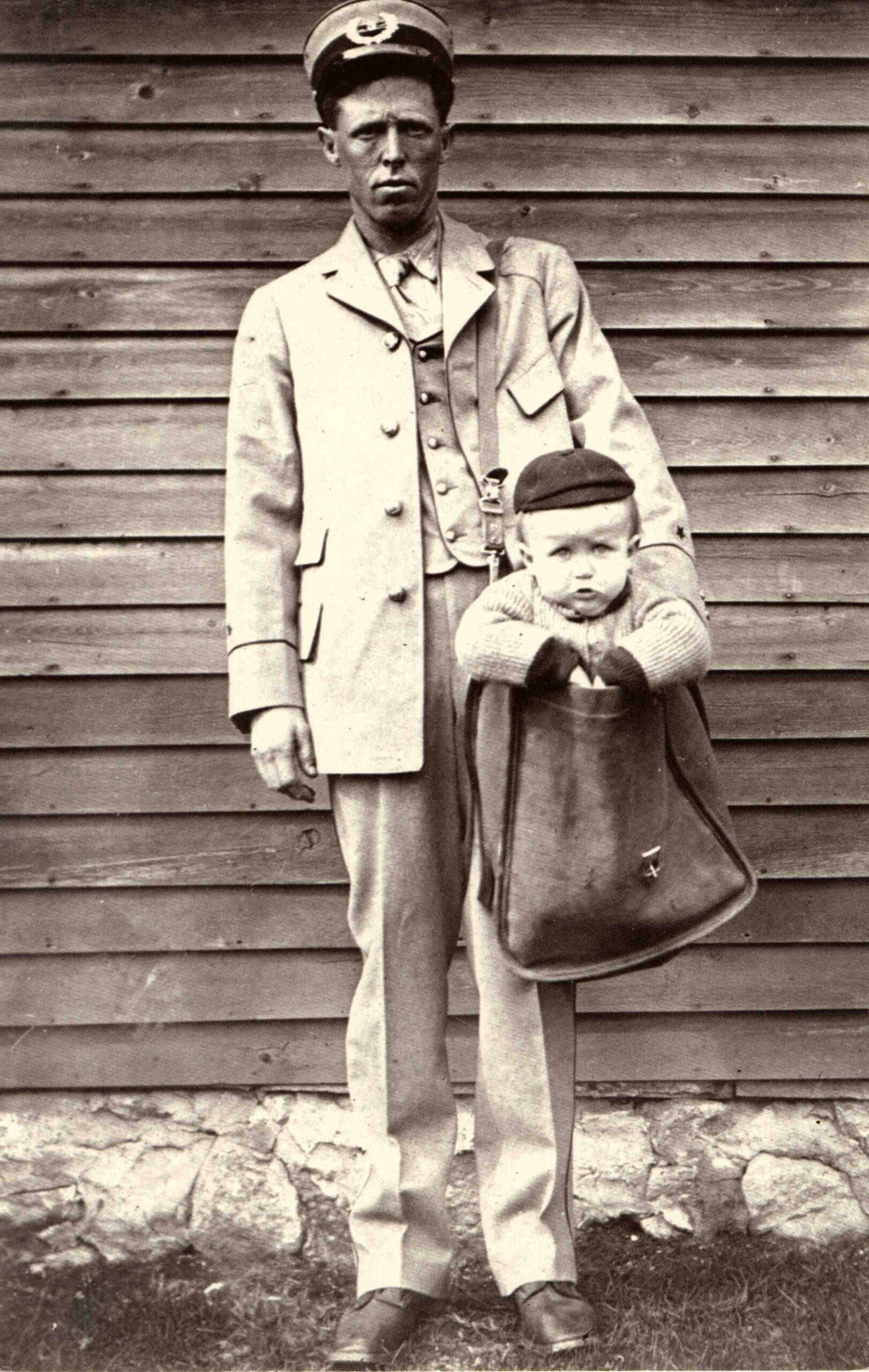 Early 1900s photo of a uniformed letter carrier posing with a little boy in a cap sitting in his leather mailbag