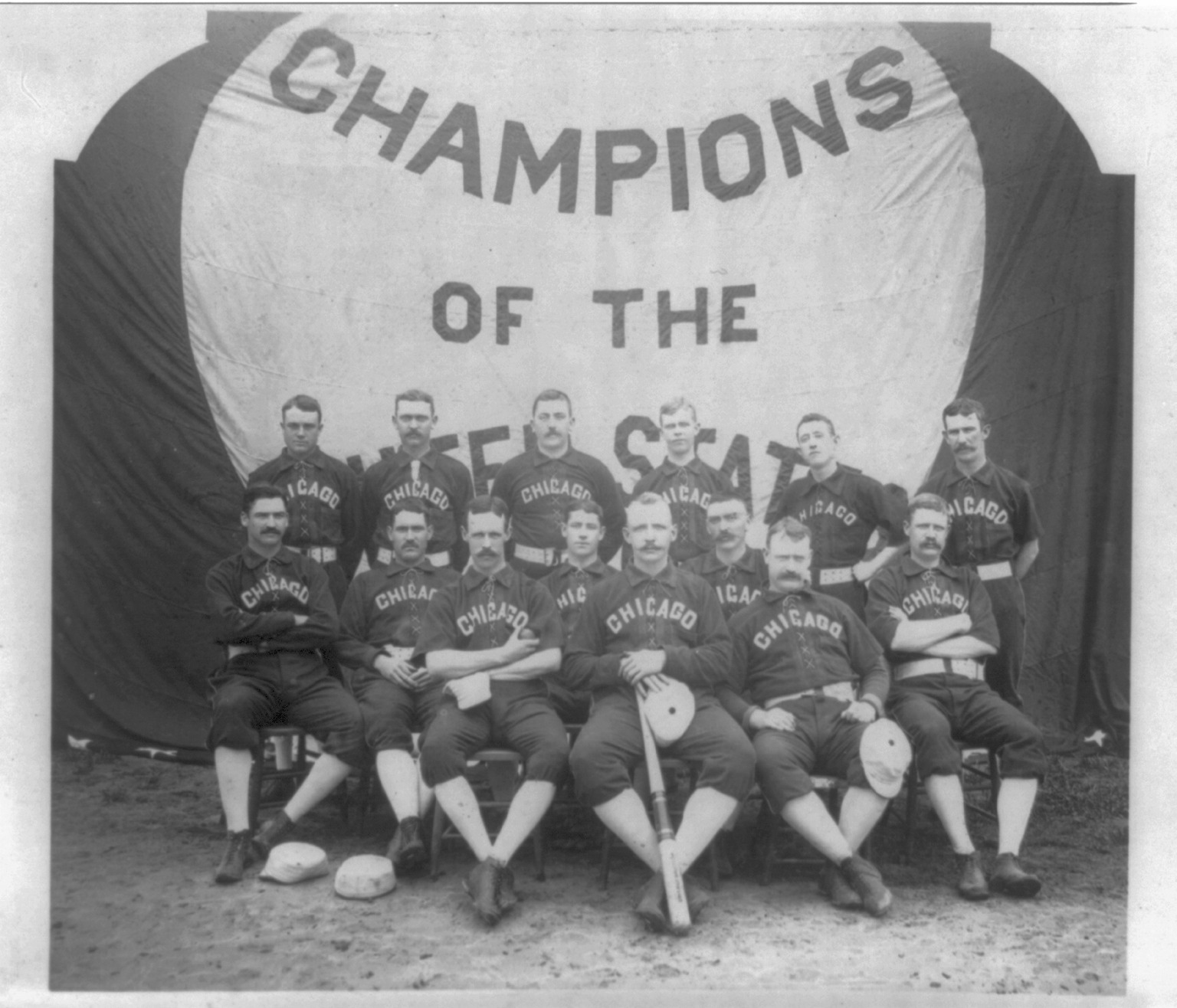 Fourteen Chicago ball players in dark uniforms lettered CHICAGO, seated and standing in front of a huge banner reading Champions of the United States, about 1886